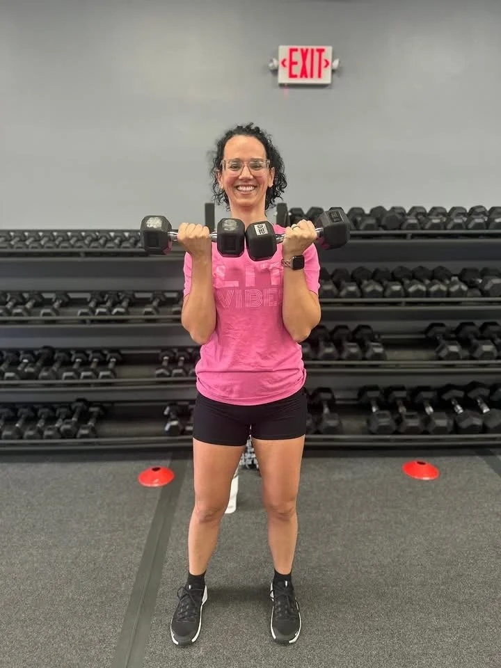 A woman smiling and holding a dumbbell at a gym, standing in front of a rack of weights and a gray wall with an exit sign above.
