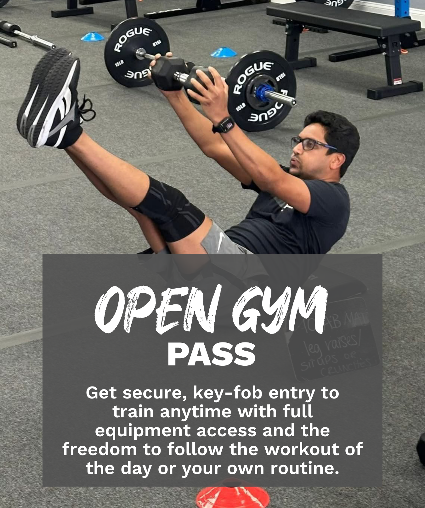 A man wearing glasses and a black t-shirt lifting a dumbbell while sitting on the gym floor surrounded by workout equipment and markers.