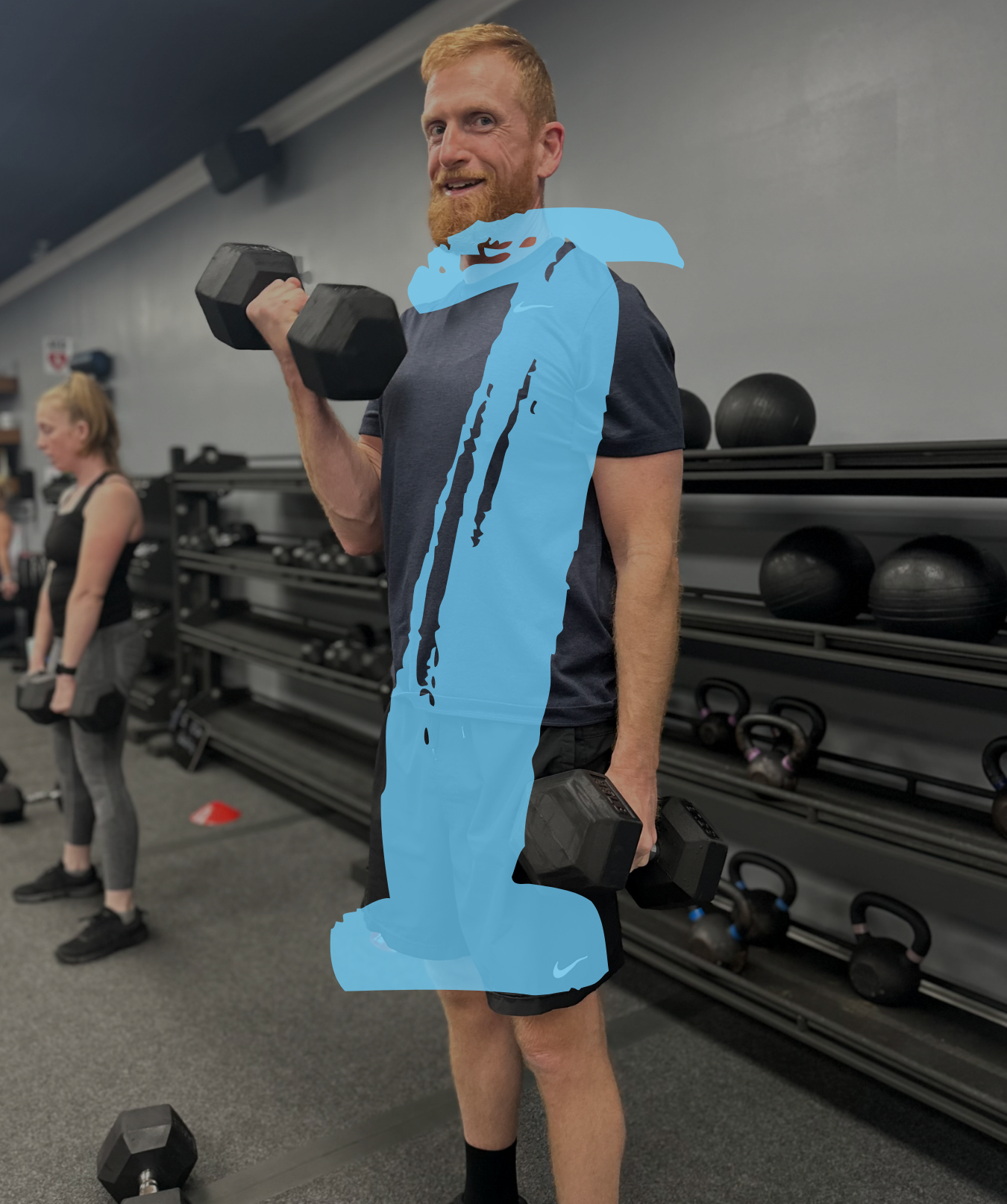 A man with a beard and red hair lifting black dumbbells at a gym, with a woman in the background also lifting dumbbells.