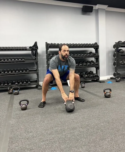 Man in workout clothes lifting a kettlebell in gym with dumbbell racks behind him.