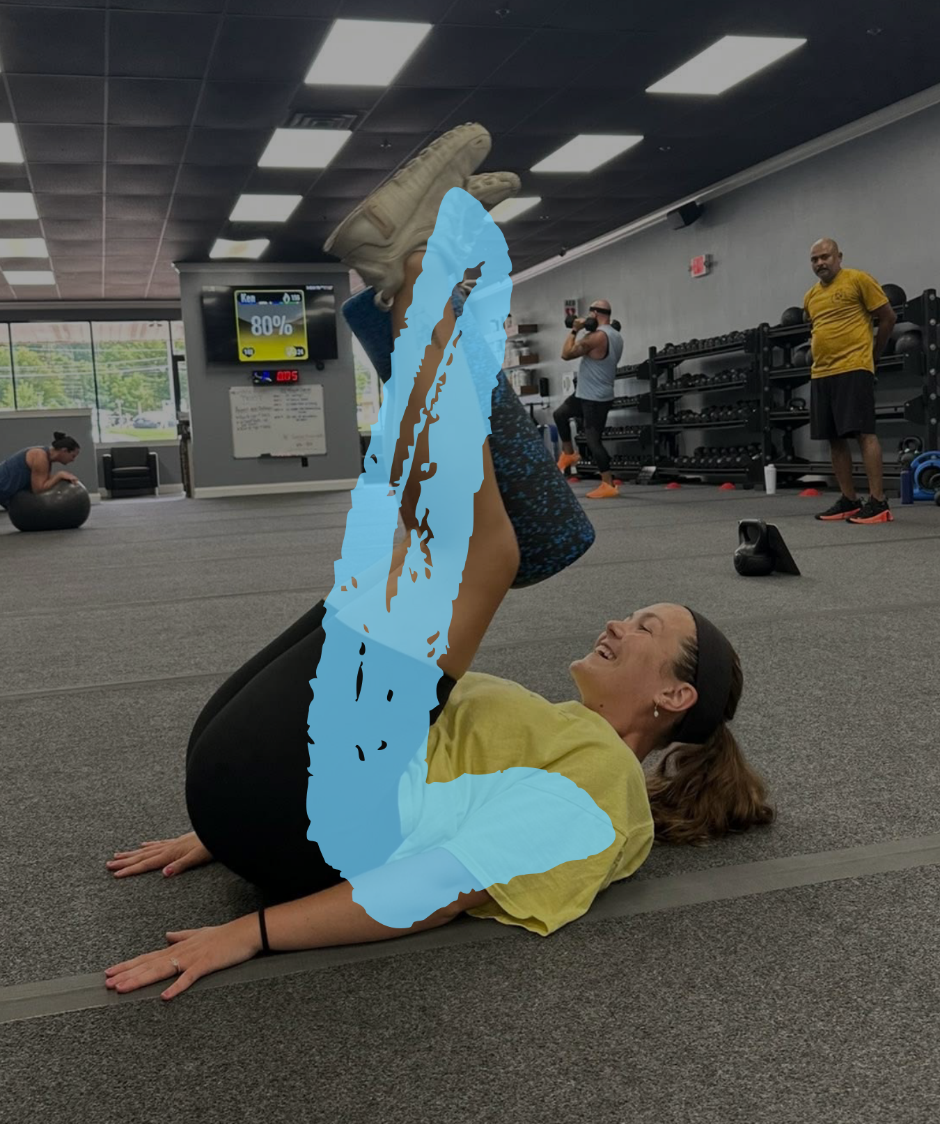 A woman performing a lying leg raise exercise in a gym, with several other people working out in the background.
