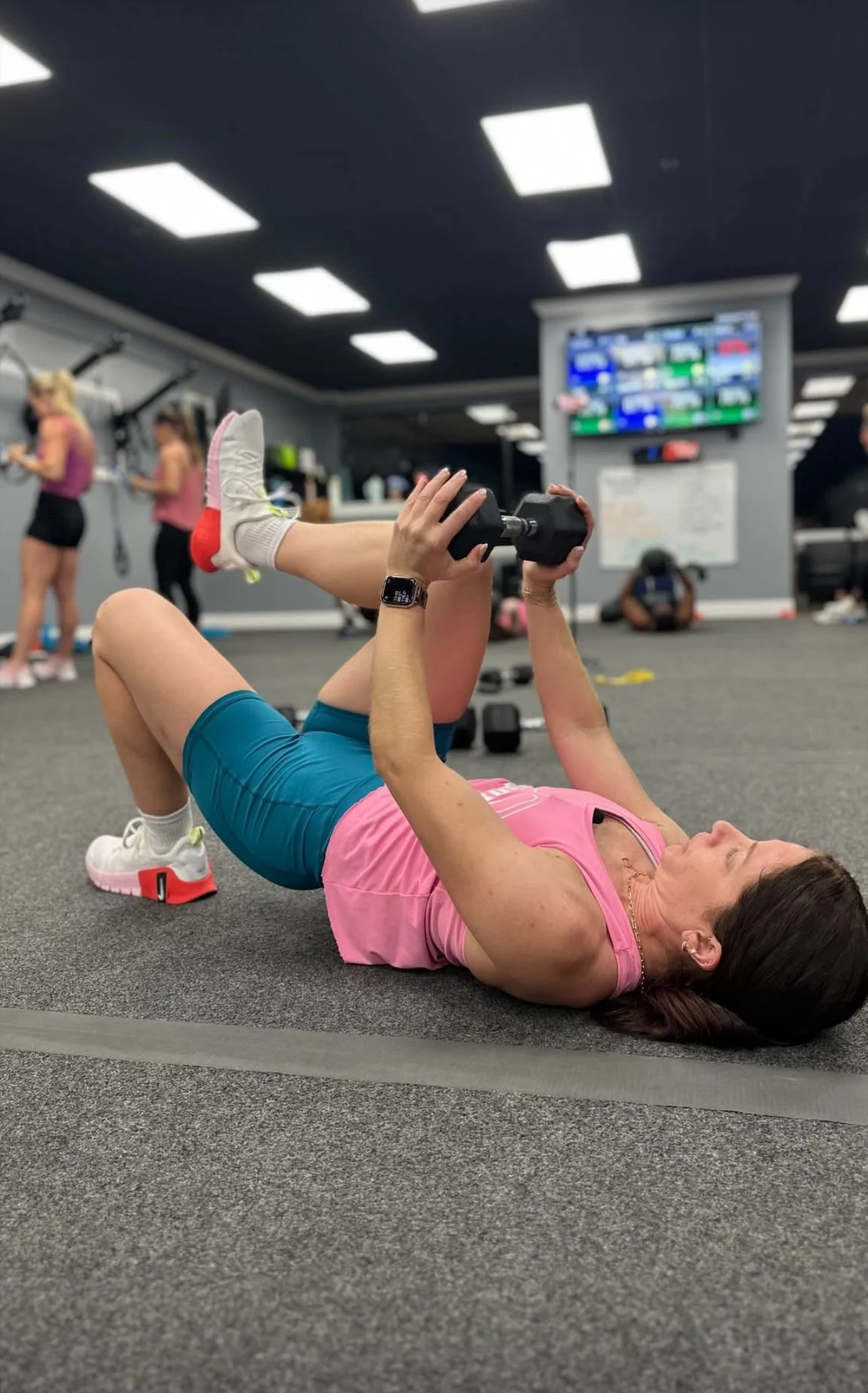 Woman lying on the gym floor lifting a dumbbell, wearing pink top and blue shorts, in a fitness center with other exercisers in the background.