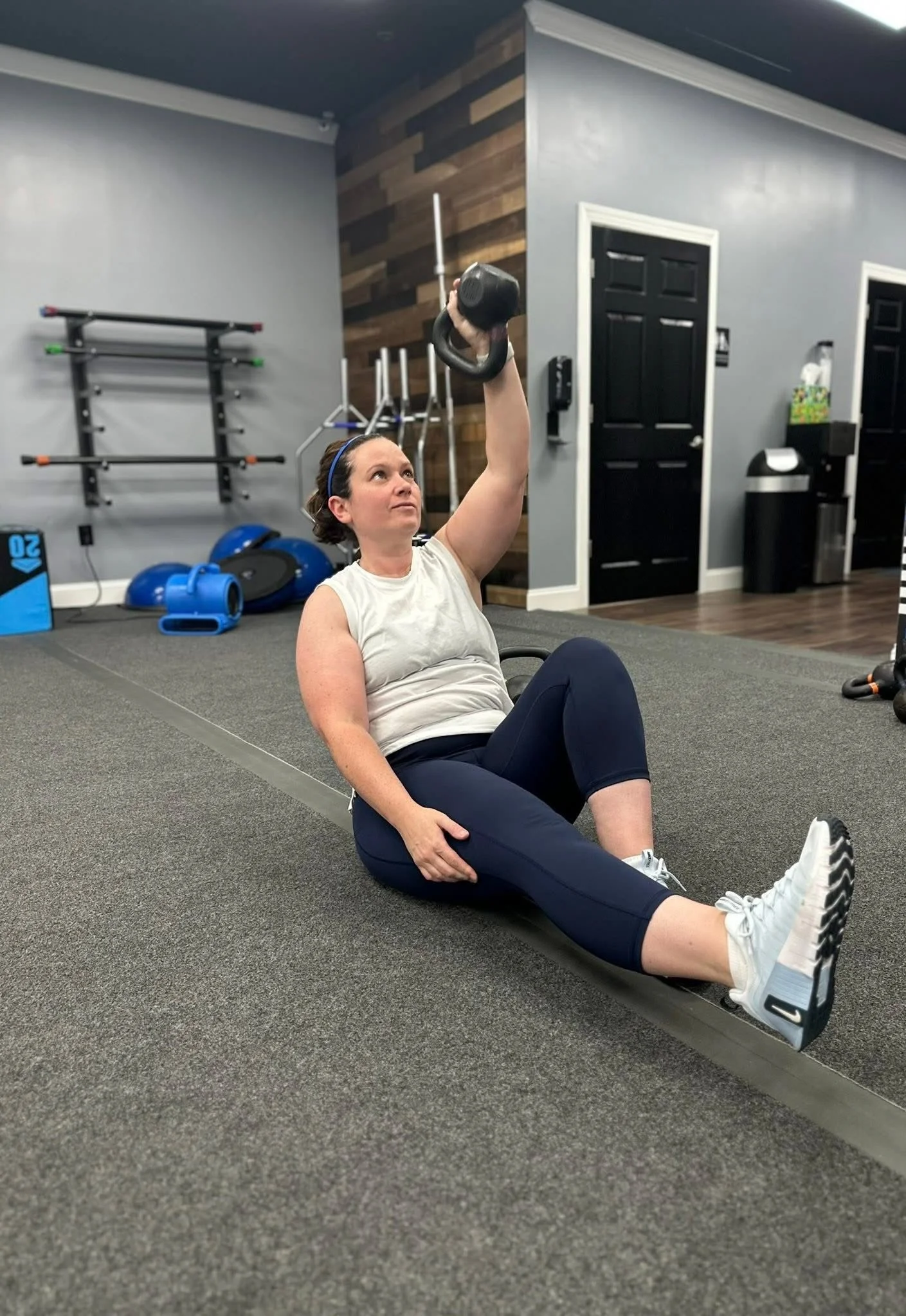 A woman sits on the gym floor holding a kettlebell overhead with her right hand while sitting with her legs extended and crossed. She is wearing athletic clothing and appears to be in a gym with various workout equipment in the background.