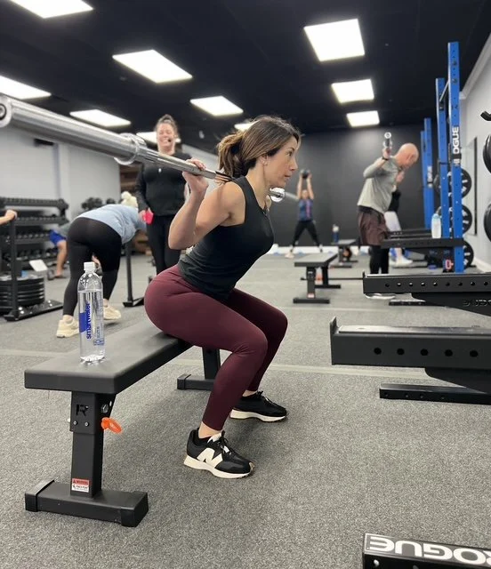 A woman doing a squat with a barbell on her shoulders in a gym, surrounded by other people exercising.