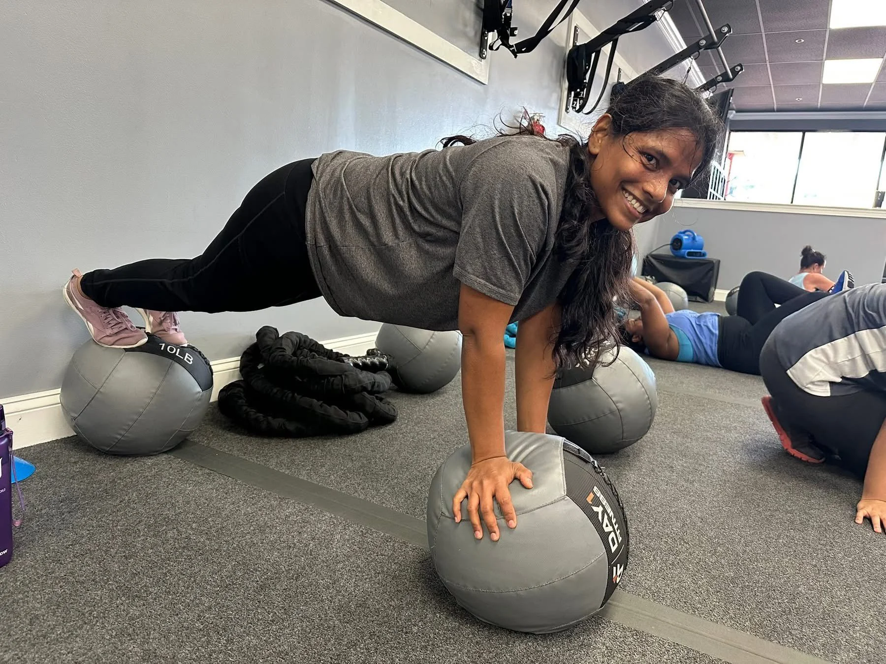Woman smiling and performing a plank exercise with her hands on a medicine ball in a gym, with other people exercising in the background.
