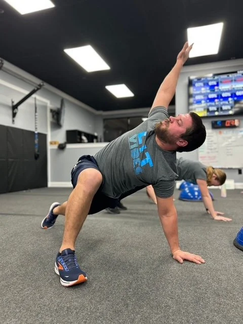 Man performing a side plank exercise in a gym, with a woman in the background.