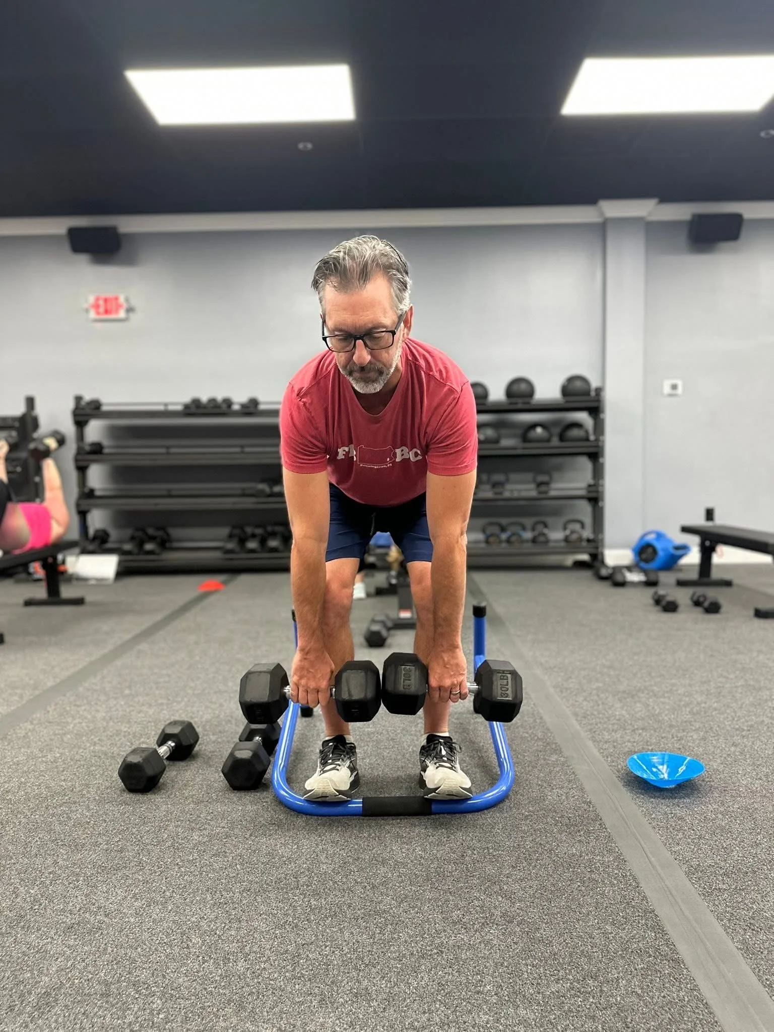 Older man with glasses and beard in a red t-shirt and dark shorts doing a push-up on a blue workout step with dumbbells in a gym.