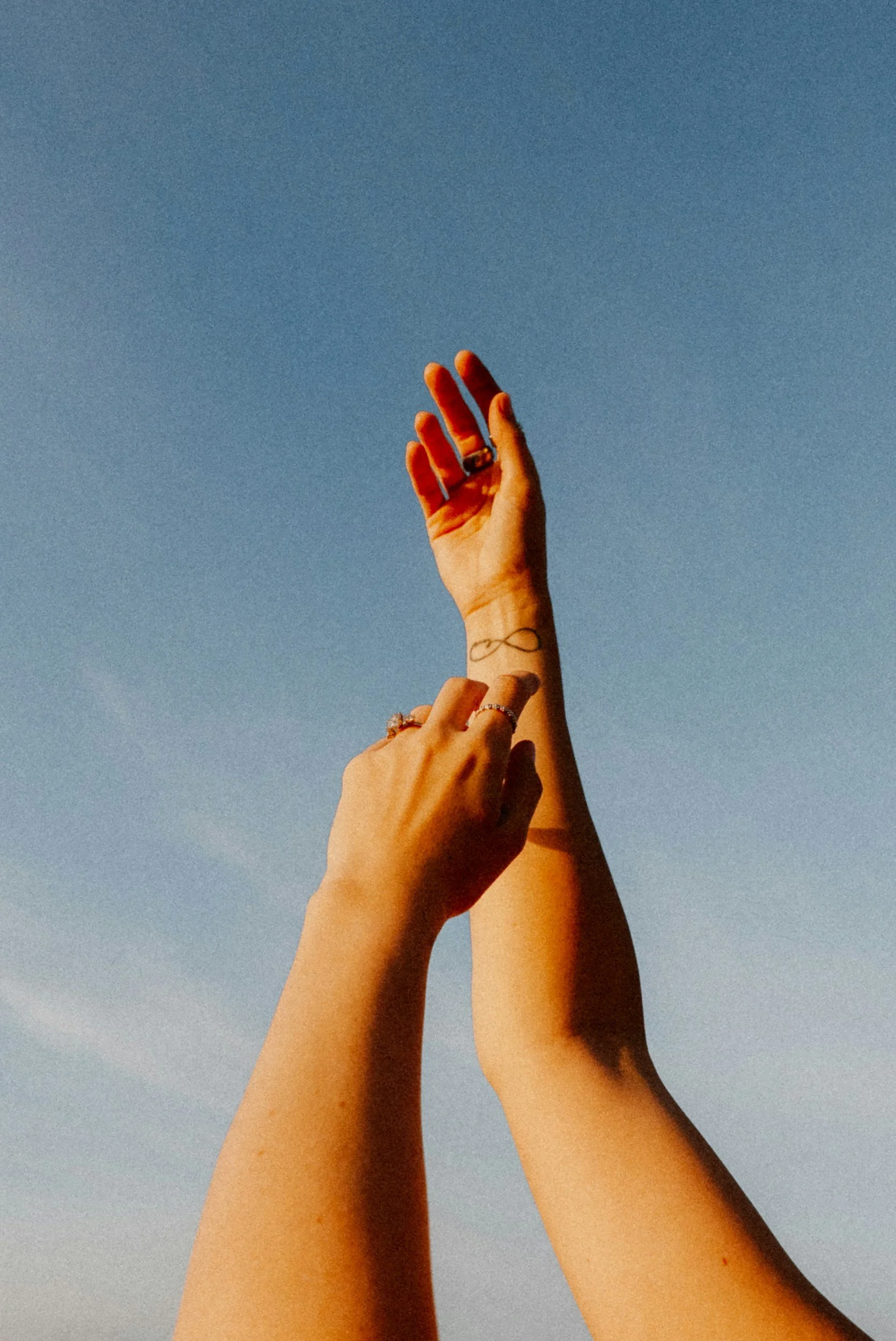 Two hands reaching upward against a clear blue sky, one hand with rings and a tattoo of an infinity symbol on the wrist.