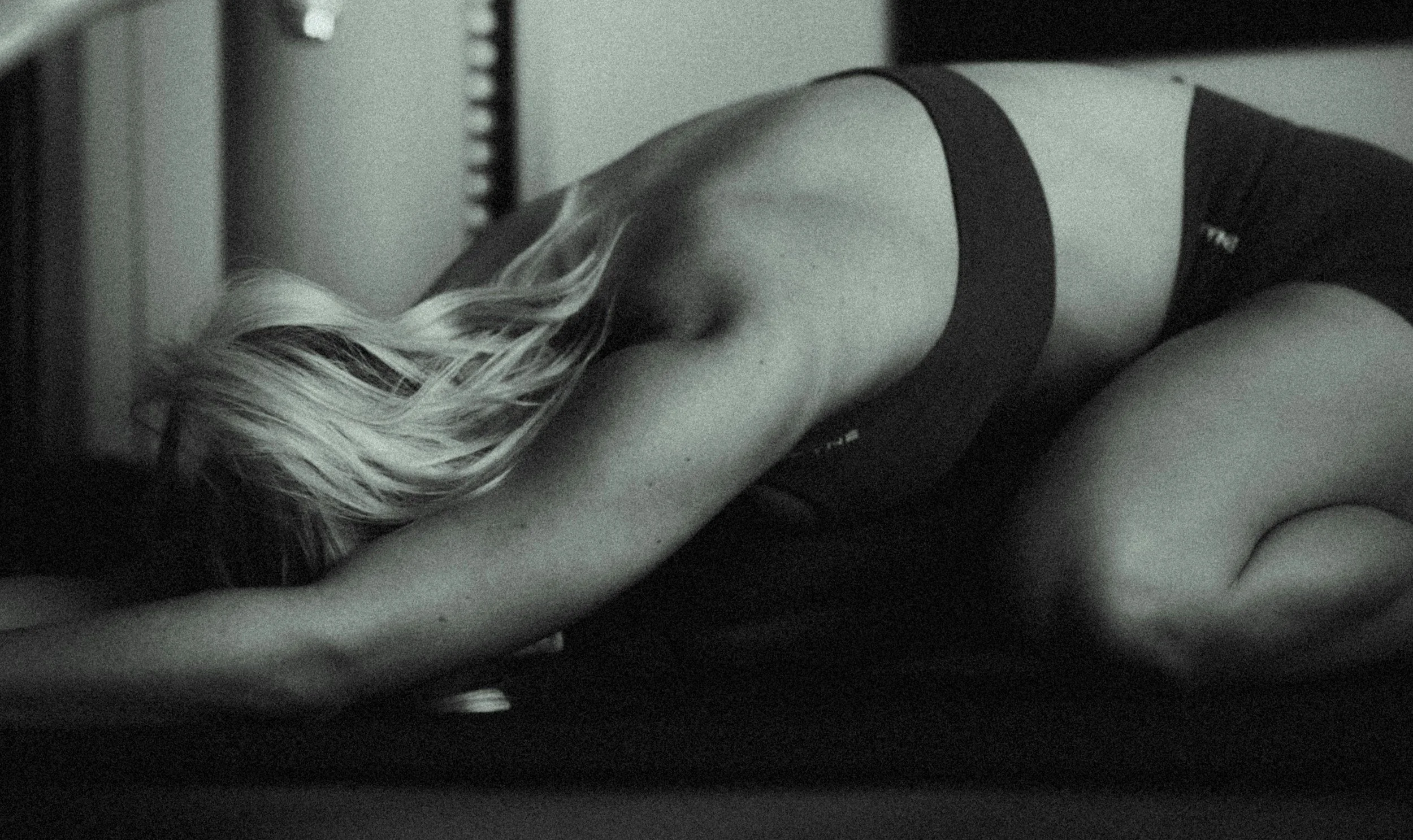 A woman with long blonde hair is bent forward on a bed, with her head and arms resting on the surface, wearing a dark sleeveless top.