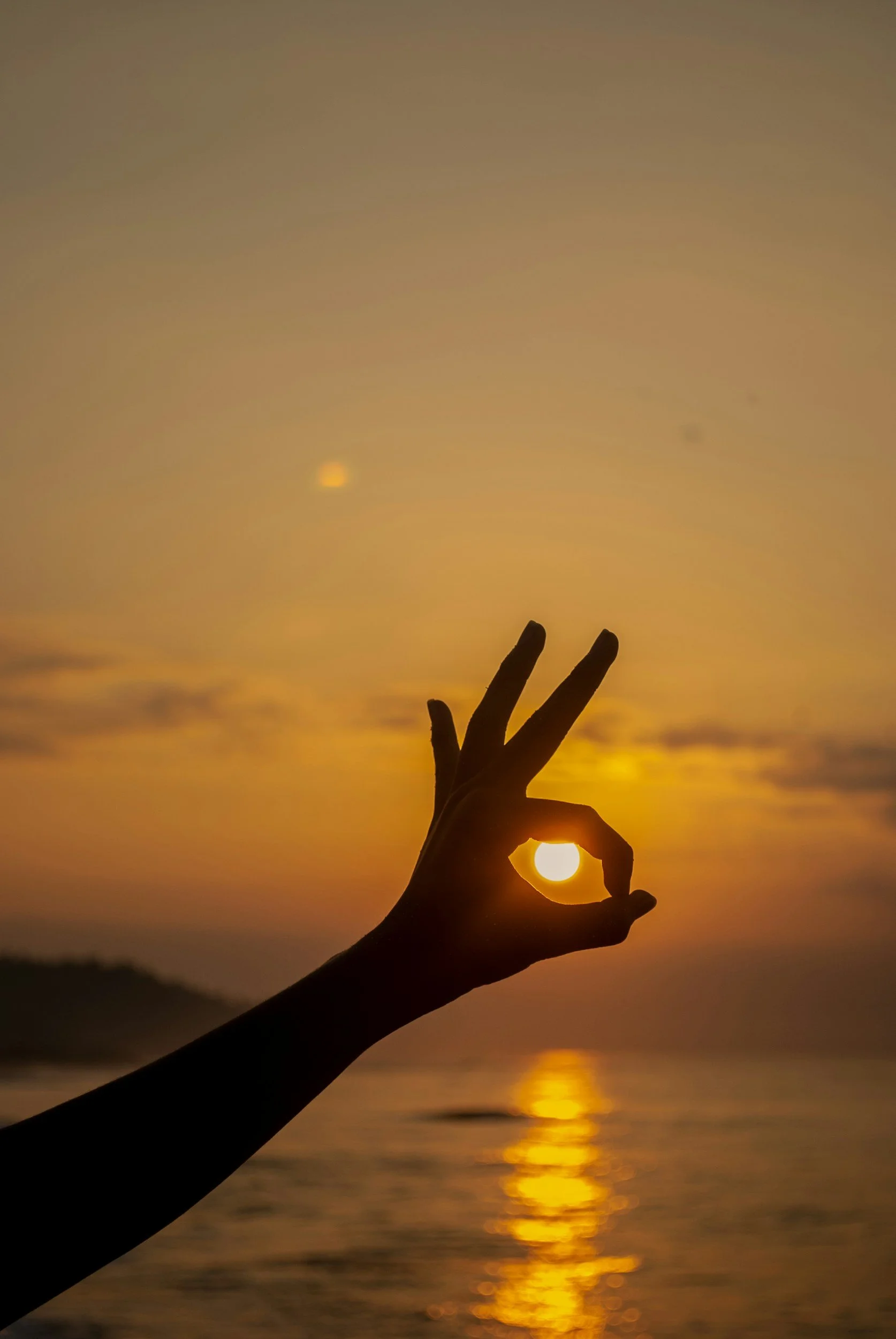 Silhouette of a hand making an 'okay' gesture with the sun aligned in the hand's circle during a sunset over water.