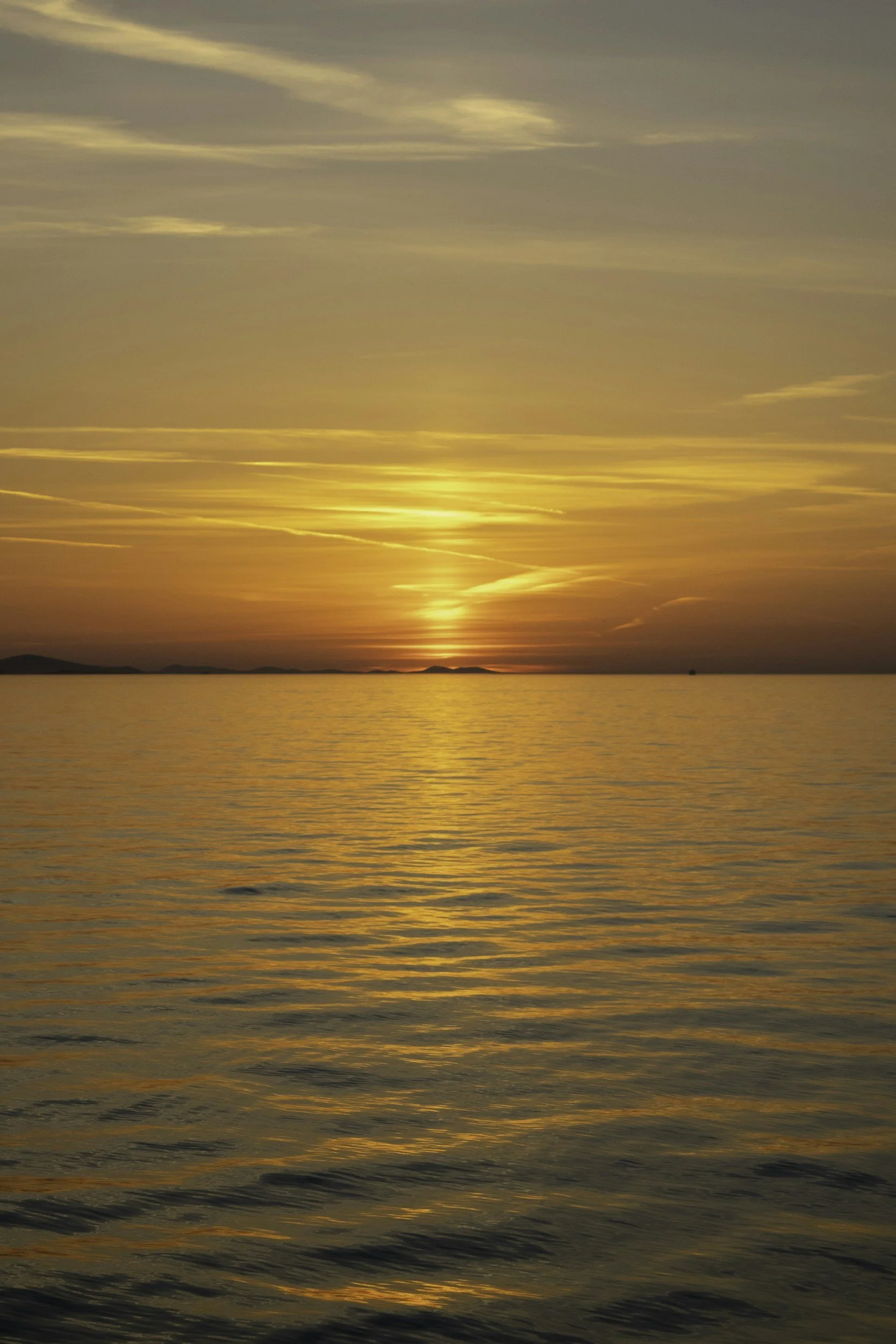 Sunset over calm water with a yellow, orange, and blue sky, and distant land on the horizon.