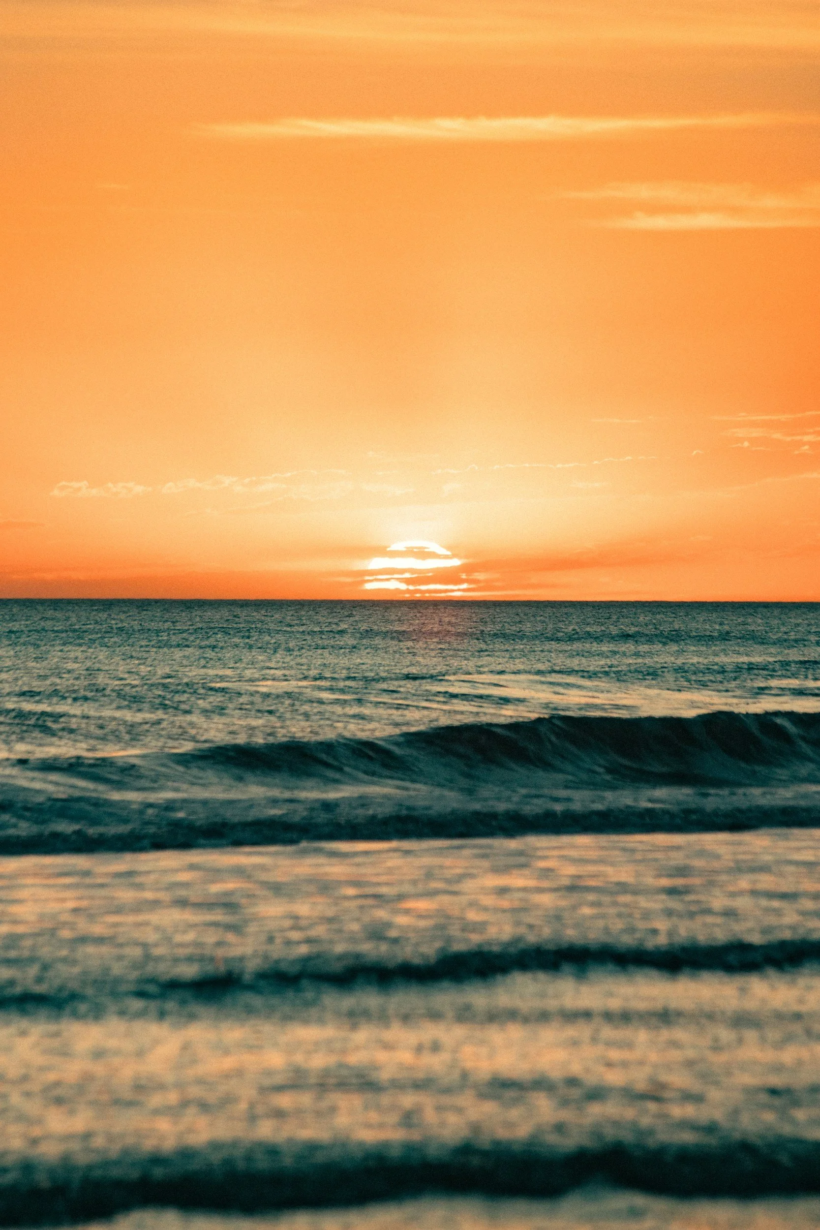 Sun setting over the ocean with orange and pink sky, waves approaching the shore.