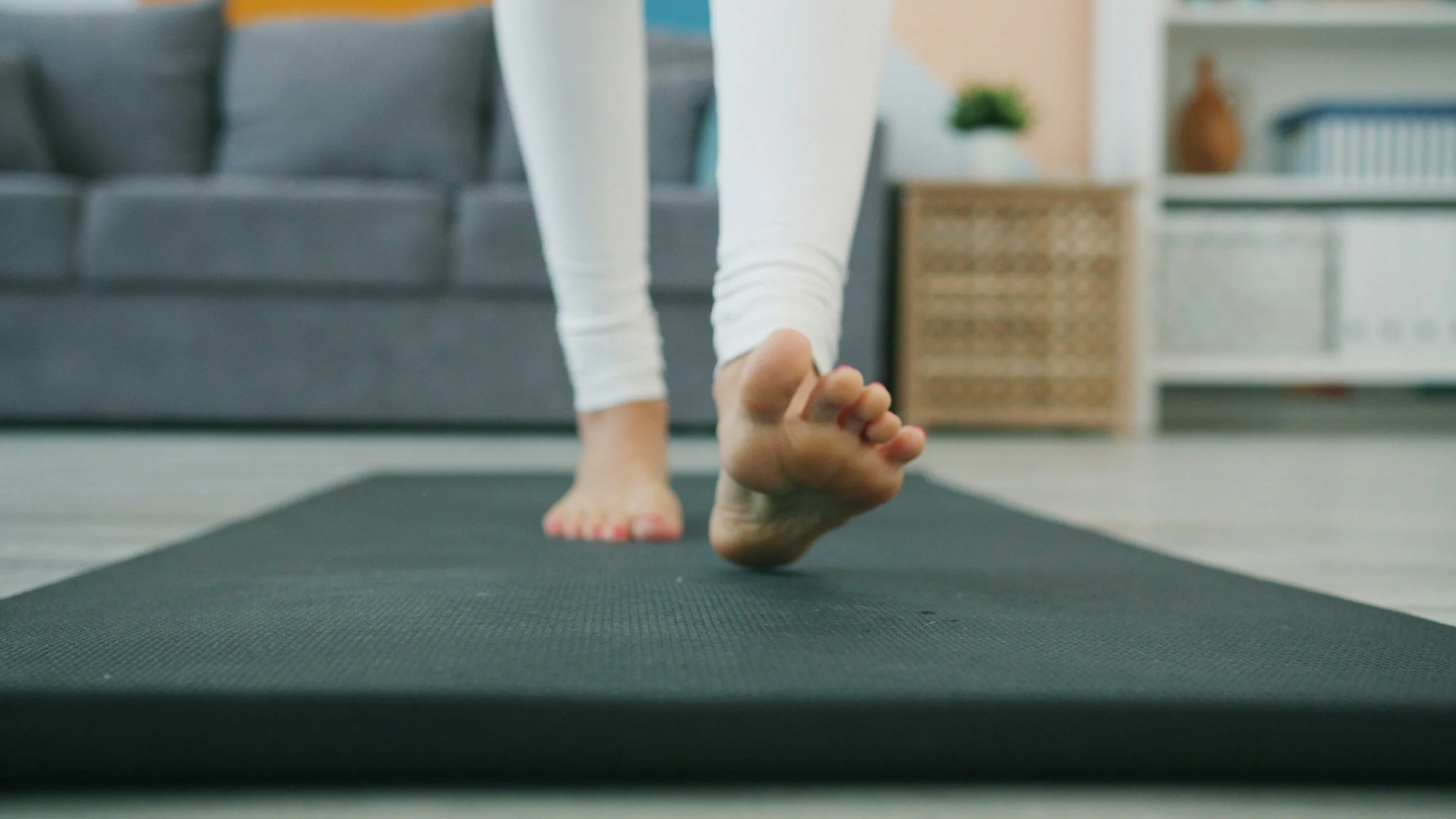 Person practicing yoga on a black mat, focusing on their feet and legs in a low lunge pose inside a living room.
