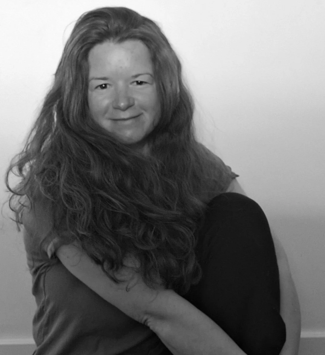 Black and white photo of a young woman with long, wavy hair, smiling, sitting on the floor with her arm around her knees.