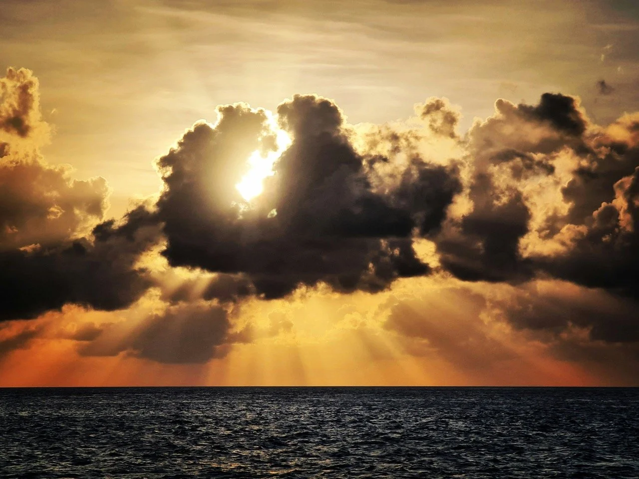 Sun partially obscured by dark clouds over the ocean, with rays of sunlight streaming through the clouds at sunset.