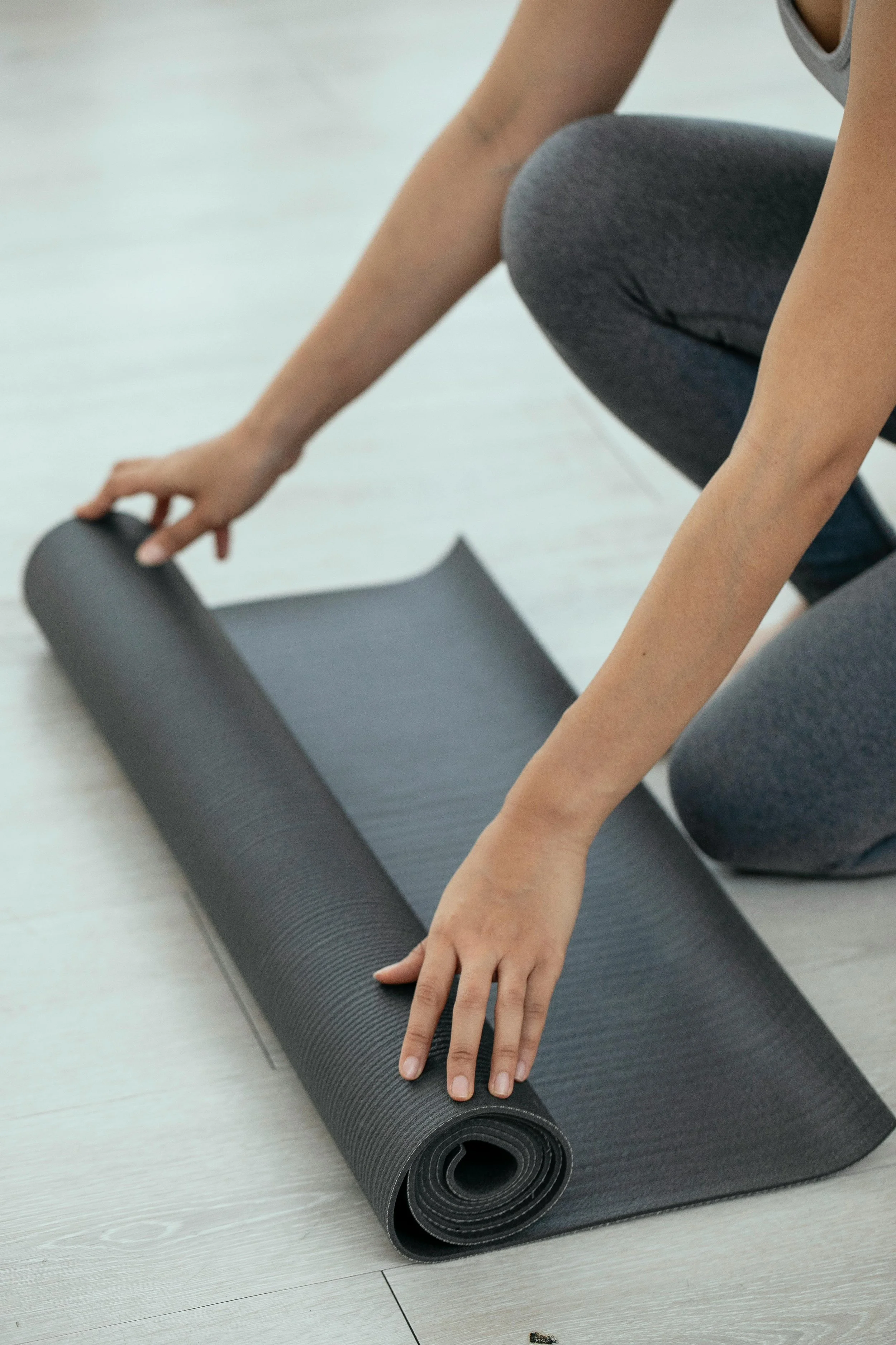Person rolling out a black exercise or yoga mat on a light-colored wooden floor.