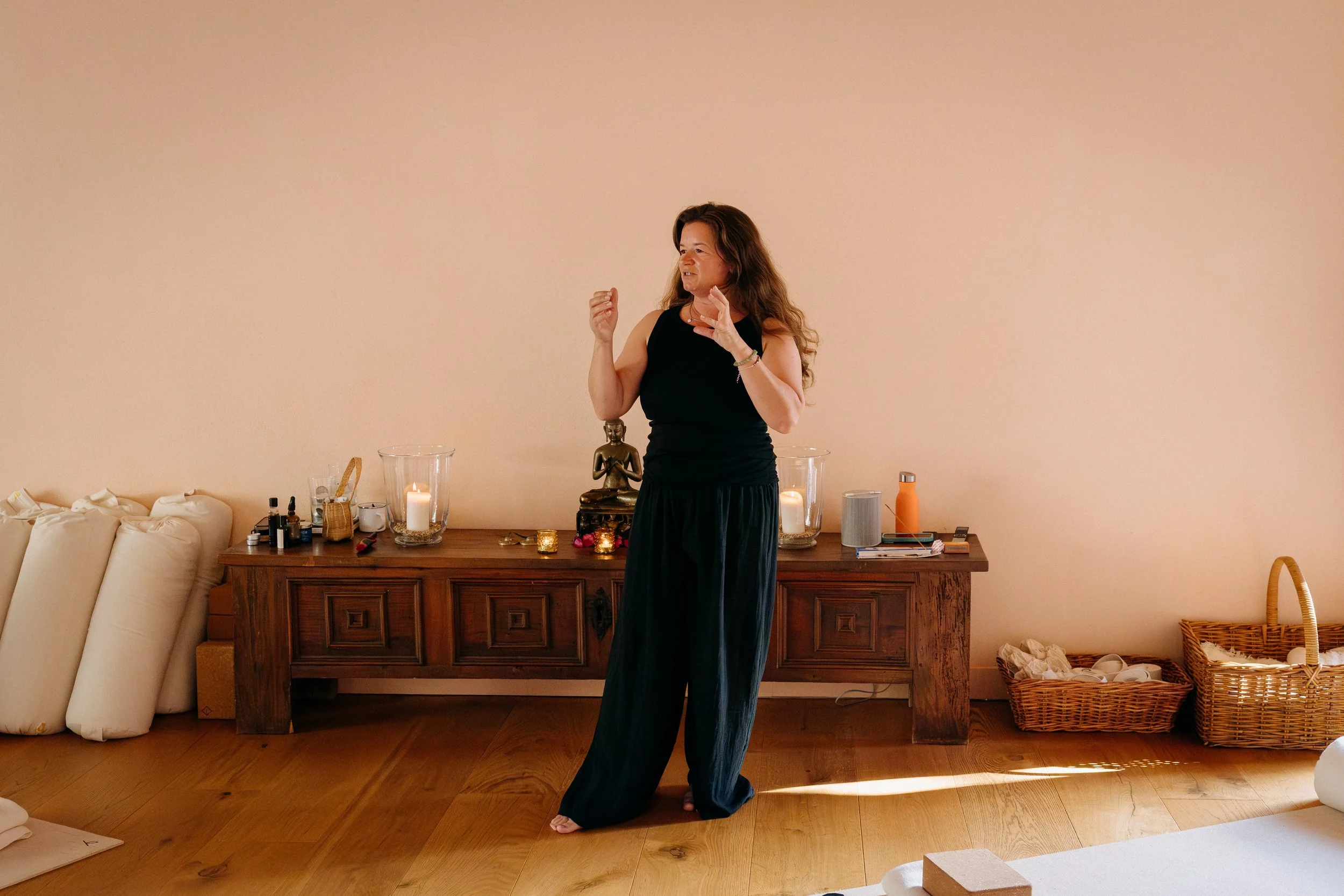 A woman in black clothing gestures with her hands in a room with a wooden floor and a long wooden sideboard decorated with candles, a small Buddha statue, and various objects. Two wicker baskets and some rolled-up cushions lean against the wall on the left and right sides of the sideboard.