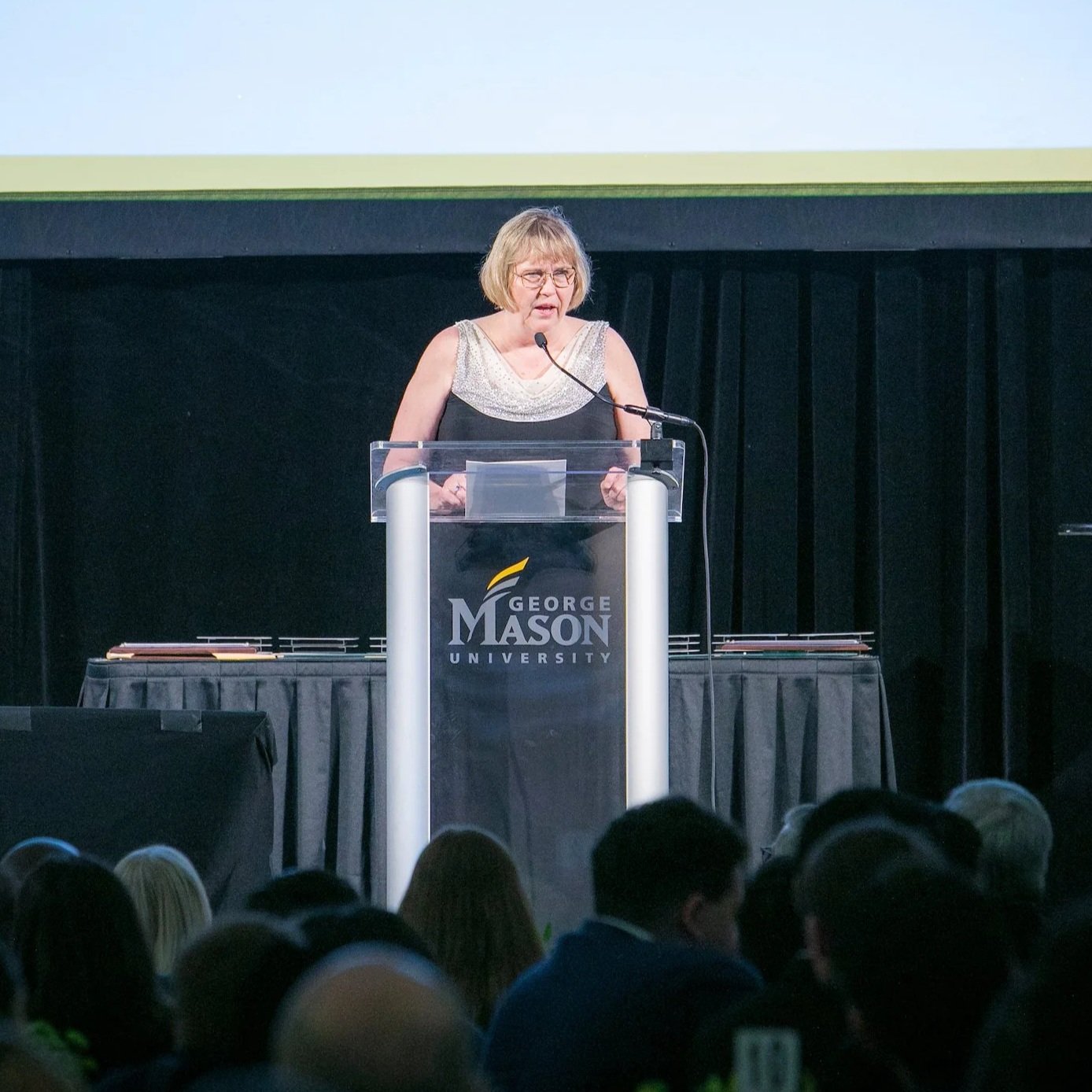 A woman standing behind a clear podium on stage, speaking into a microphone during a presentation at George Mason University, with an audience in front of her.