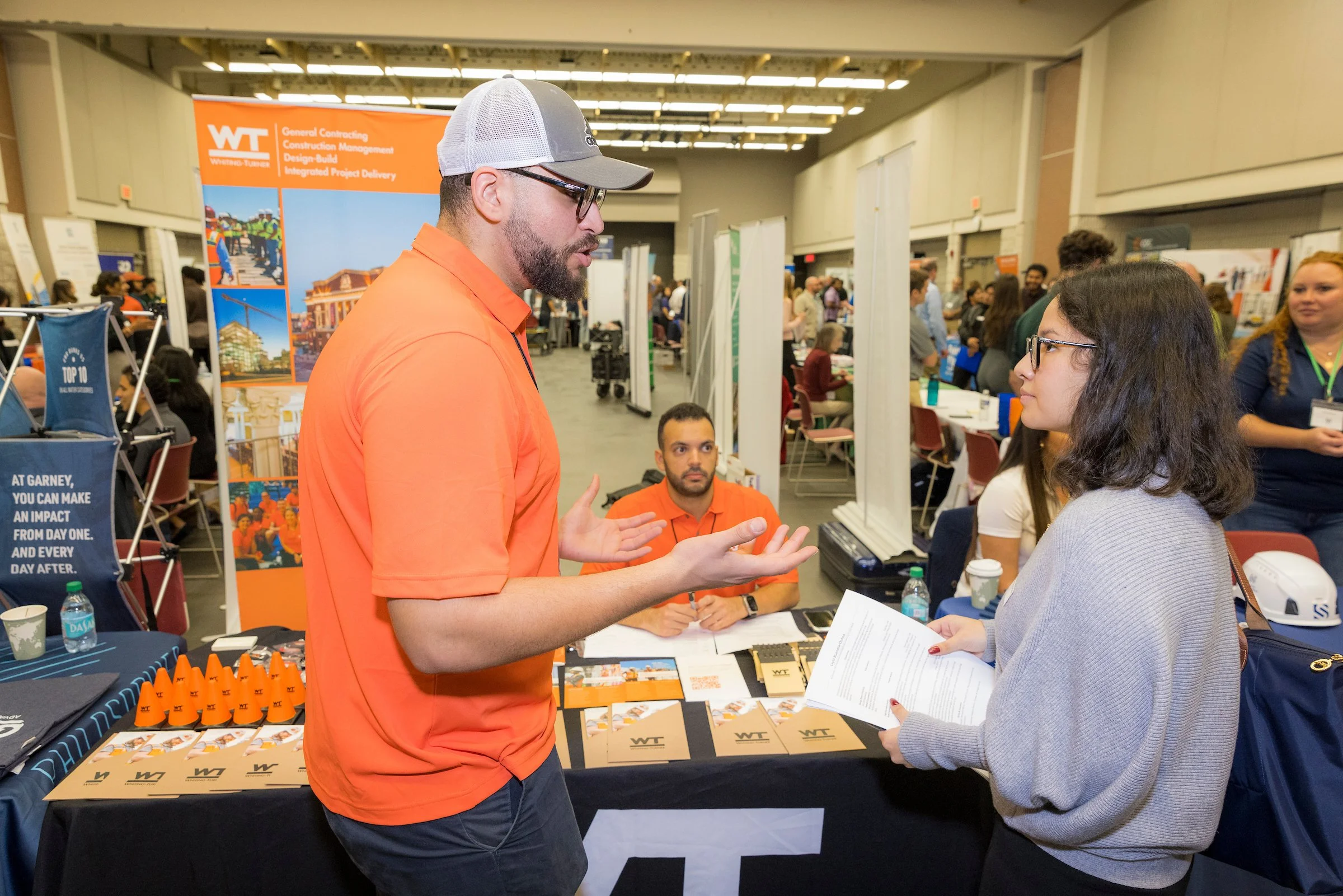 A man and a woman are engaged in a conversation at a career or job fair. The man is wearing an orange shirt and a gray cap, while the woman is holding a pamphlet or brochure. They are standing in front of a booth with promotional items, brochures, an