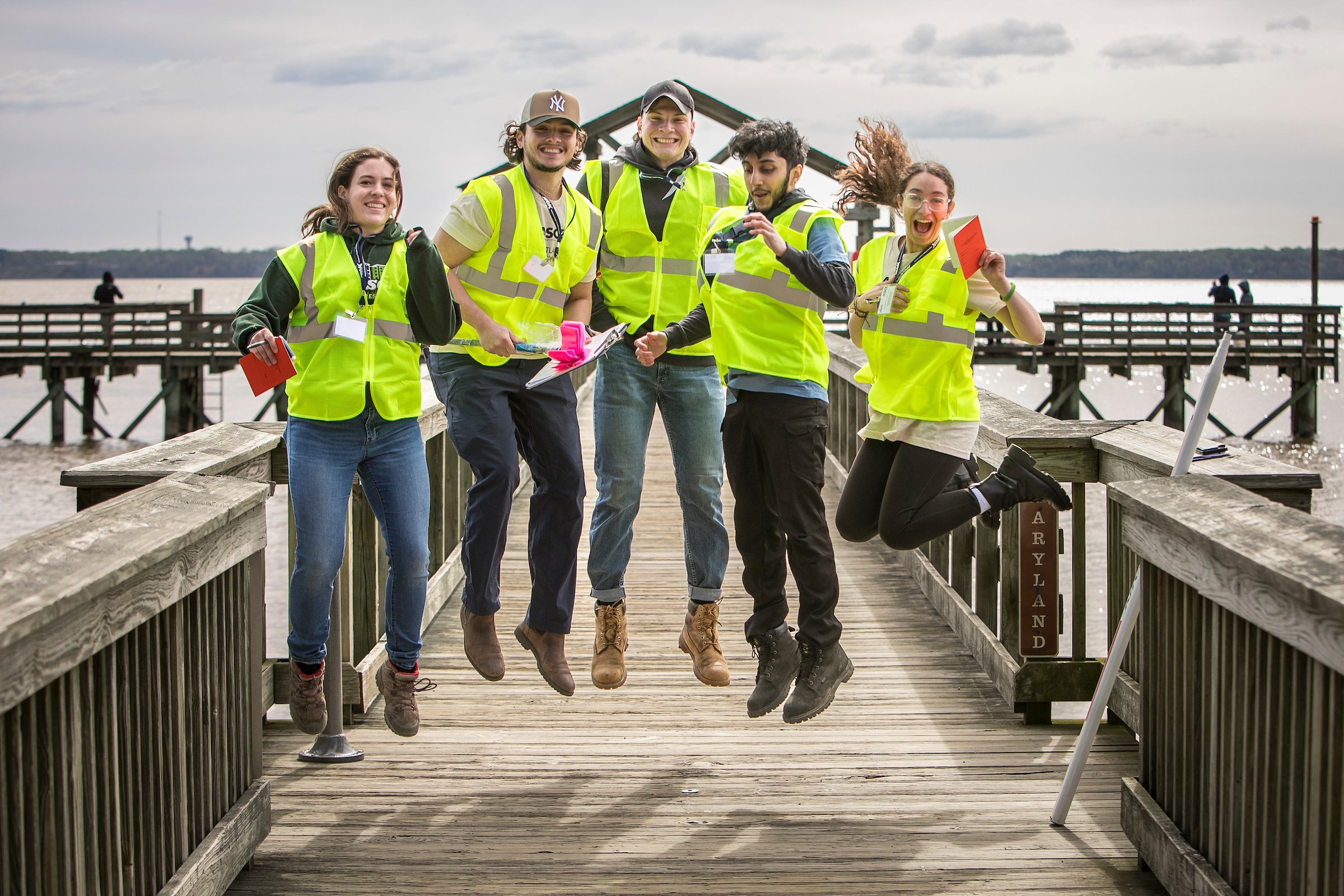 Group of five young adults wearing safety vests jumping on a wooden dock by the water, with a cloudy sky and a pier in the background.