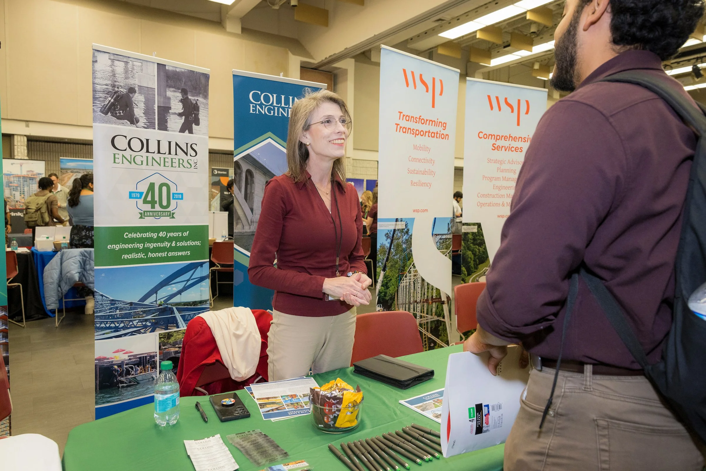 A woman in a red jacket talking to a man at an engineering conference booth. The booth has banners related to Collins Engineers and transportation services.