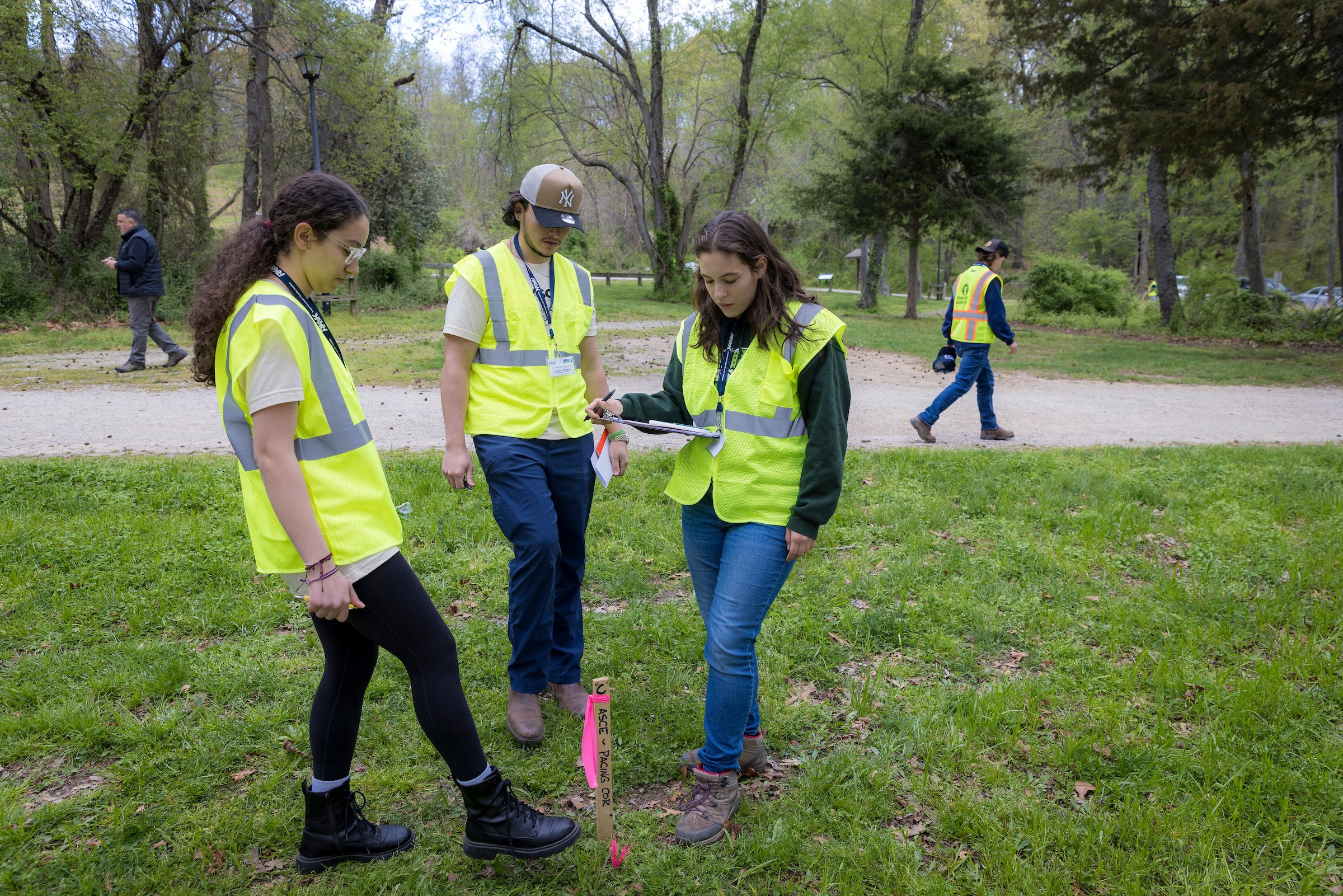Group of people in yellow vests working outdoors in a park, with some holding clipboards and notes, and a pink marker labeled 'Acre - Paving OK' in the grass.