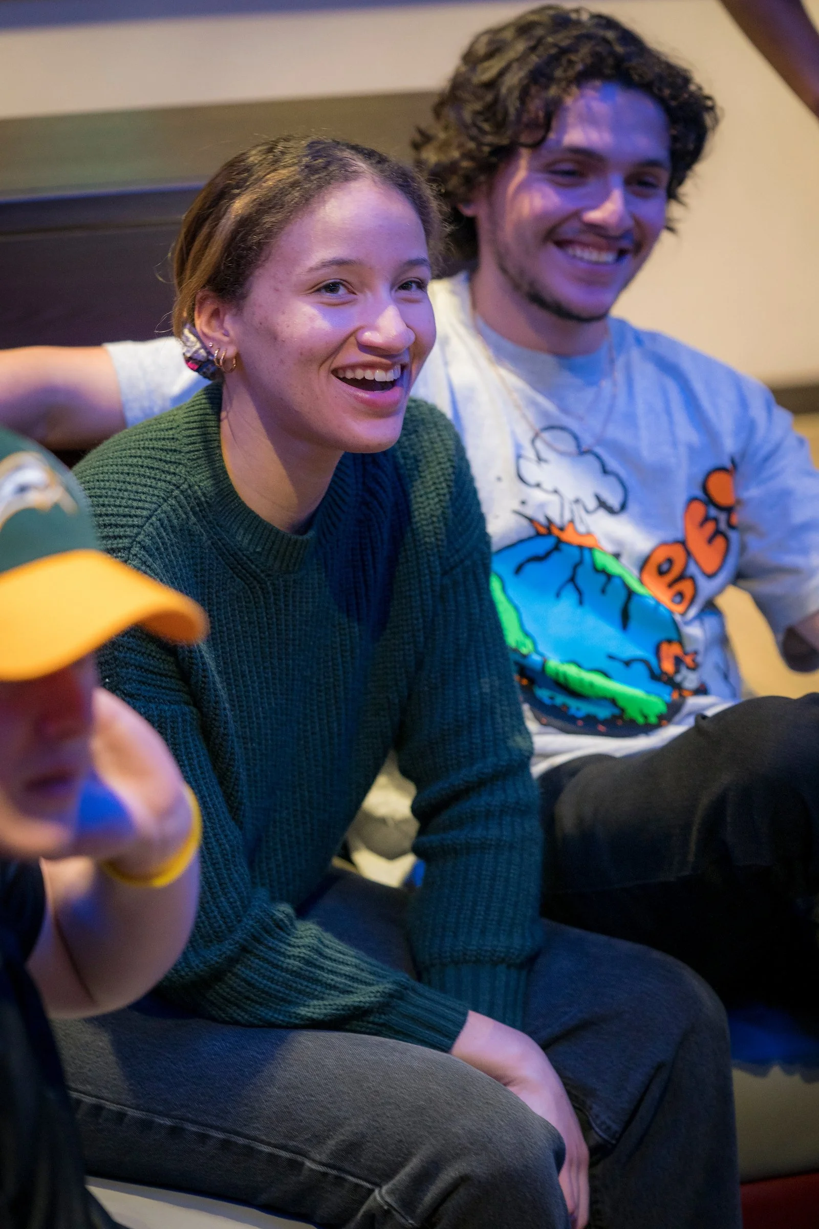 Two young adults, a woman with brown hair and a man with curly hair, smiling and sitting on a couch, with a blurred person in a green baseball cap in the foreground.