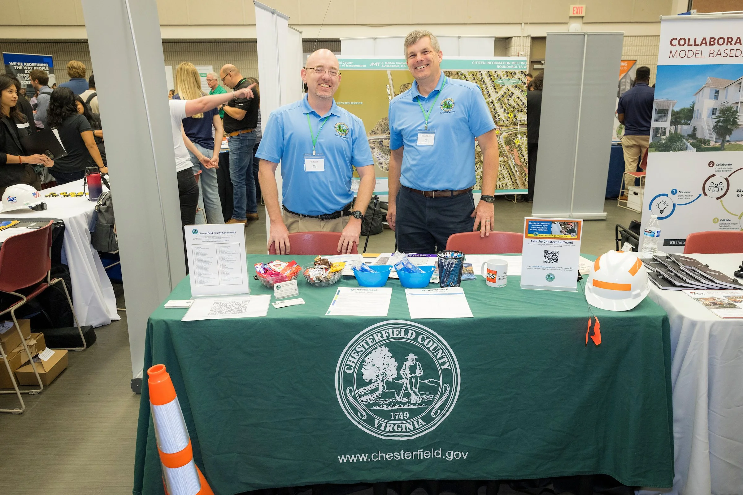 Two men standing behind a Chesterfield County, Virginia table at a conference or expo, smiling and looking at the camera. The table is covered with promotional materials, snacks, and a hard hat, with a traffic cone on the front. People and display booths are visible in the background.