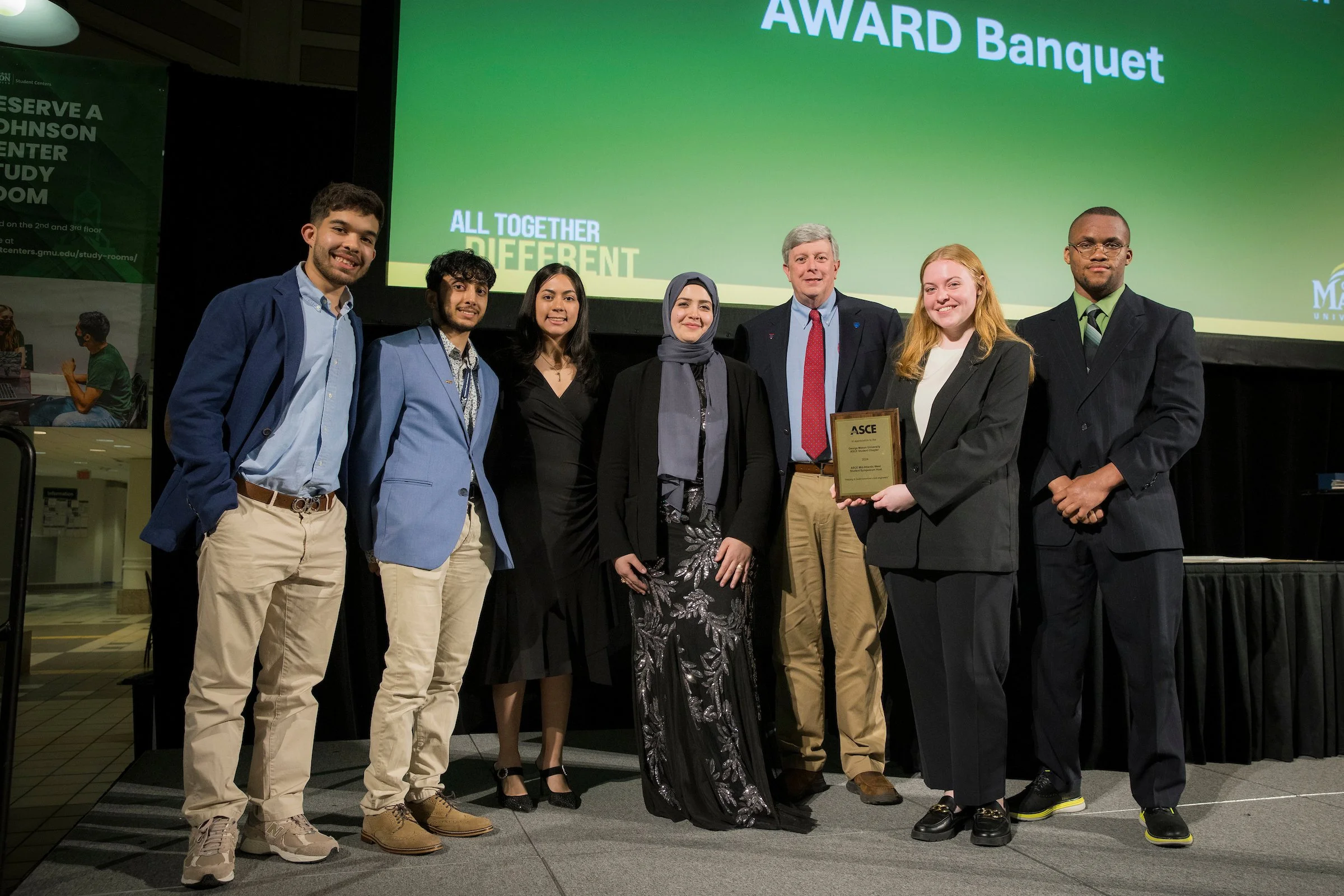 Group of seven people standing on stage at an award ceremony, with a large green screen behind them displaying the words 'AWARD Banquet' and 'ALL TOGETHER DIFFERENT'. One woman in the group is holding a framed award plaque. The group is smiling and dressed in business or semi-formal attire.