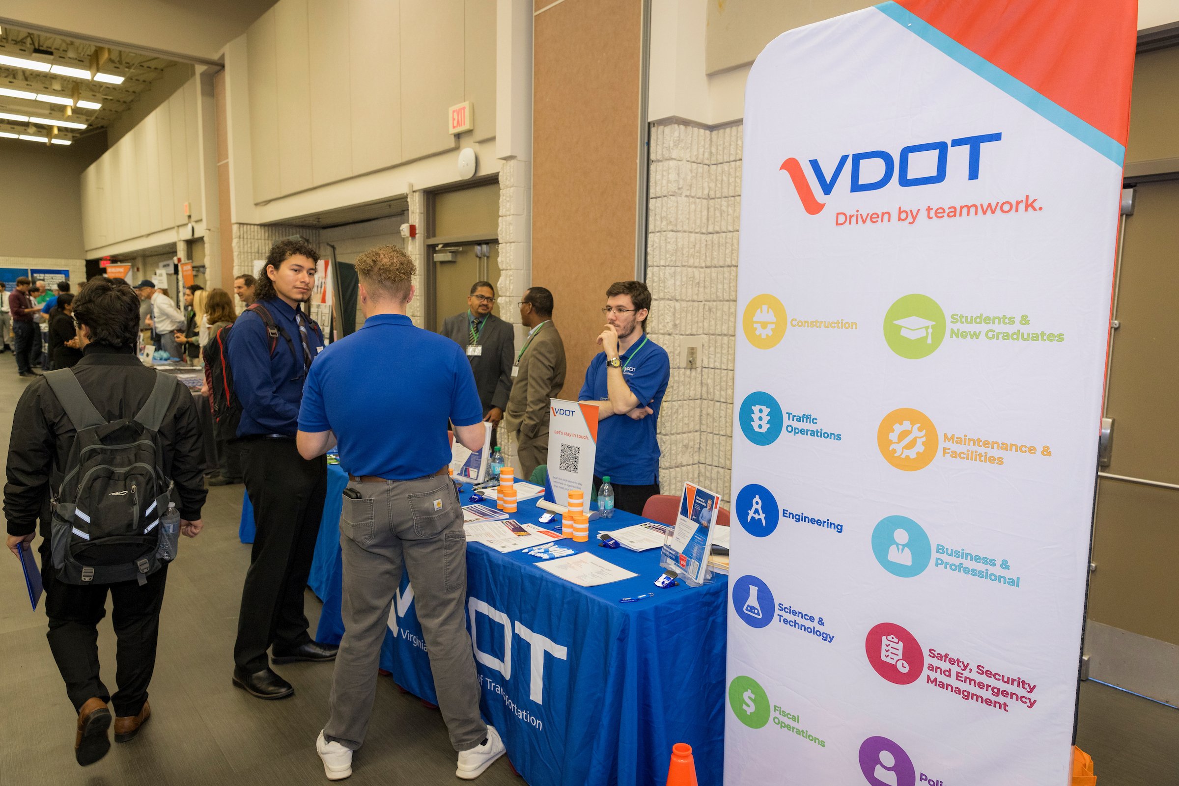 People attending a career fair or conference at a Virginia Department of Transportation (VDOT) booth. The booth has a large vertical banner displaying VDOT logo and various categories like construction, engineering, safety, and more.