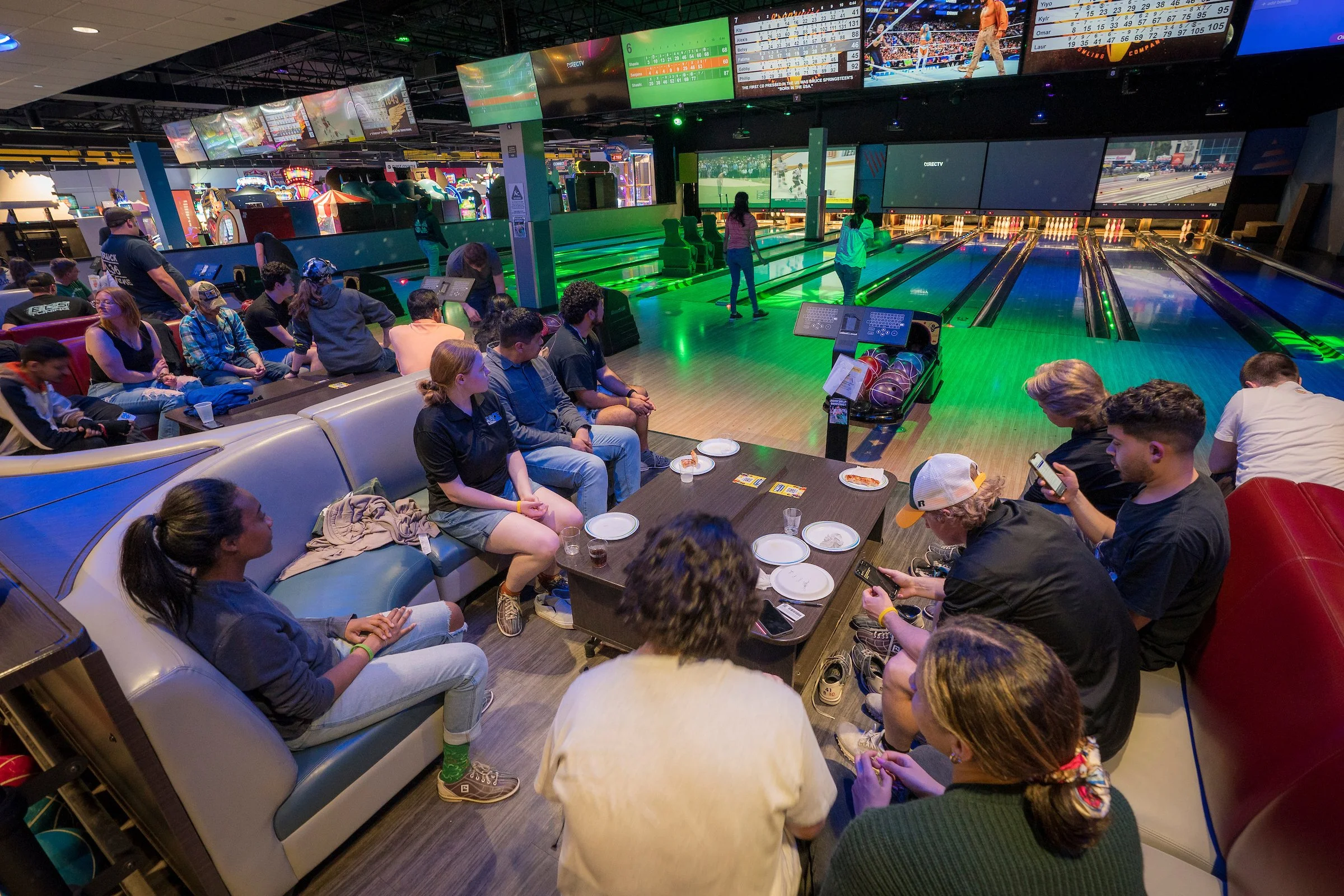 People sitting on couches and chairs watching a bowling game in a lively indoor bowling alley, with several people walking around and playing games in the background.