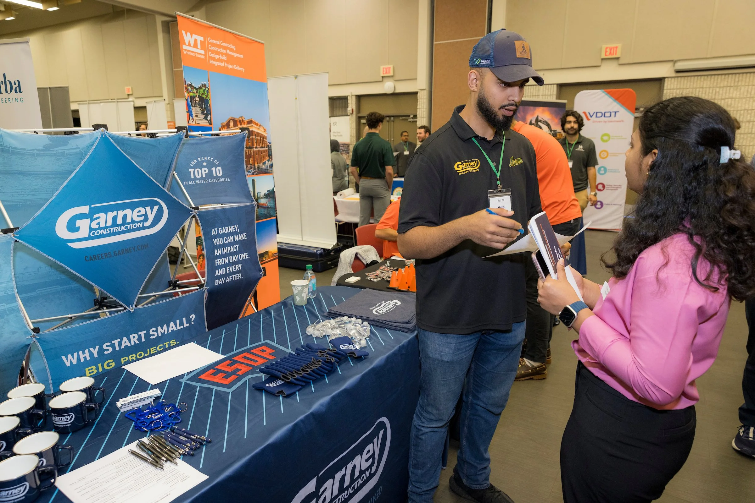 A man and woman are talking at a trade show booth for Garney Construction, with promotional merchandise like mugs, pens, and lanyards on the table.
