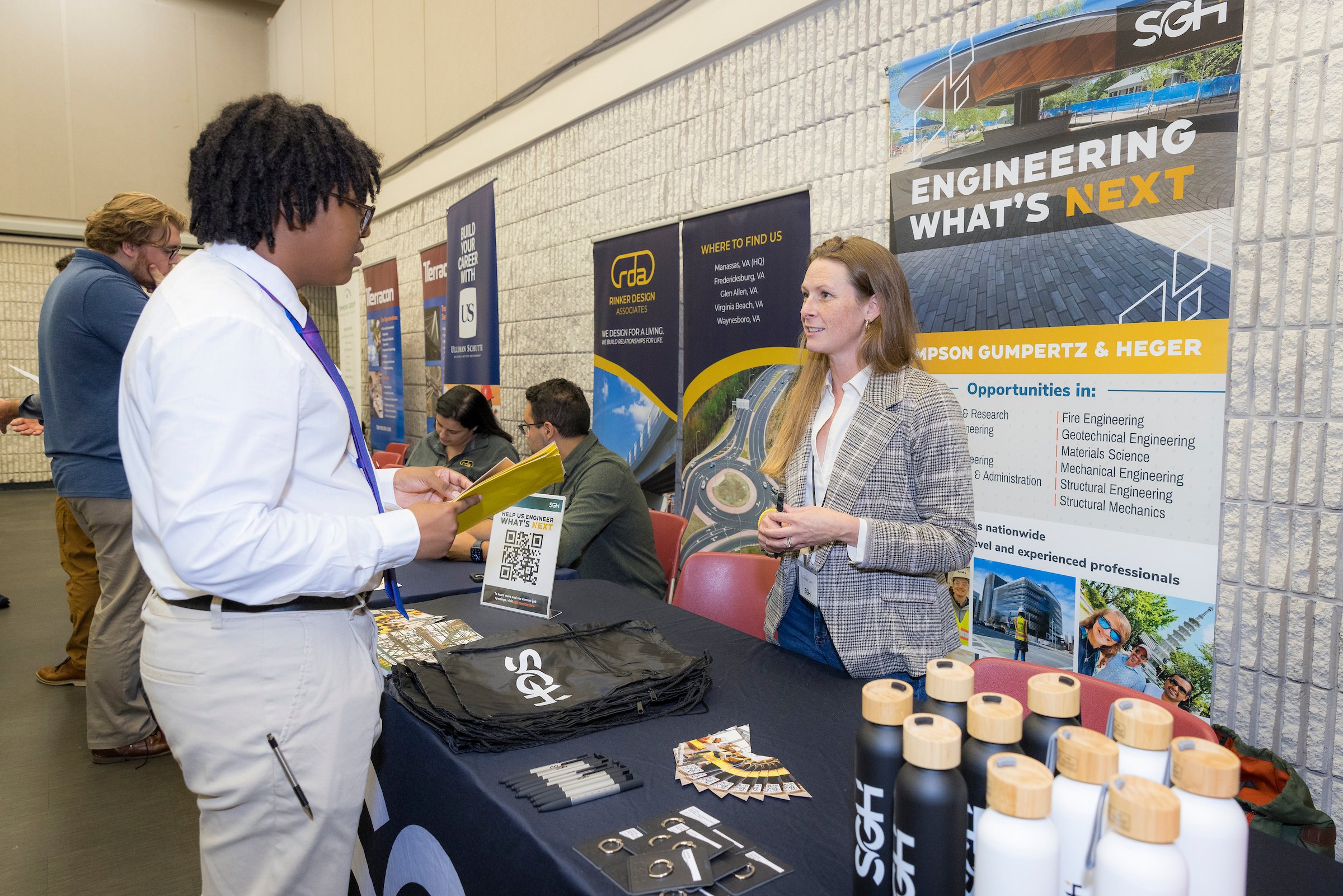 A career fair booth for engineering opportunities, with a woman in a gray plaid blazer talking to a young man in a white shirt and glasses. The booth has a black table with pens, water bottles, and pamphlets, and posters about engineering fields and 
