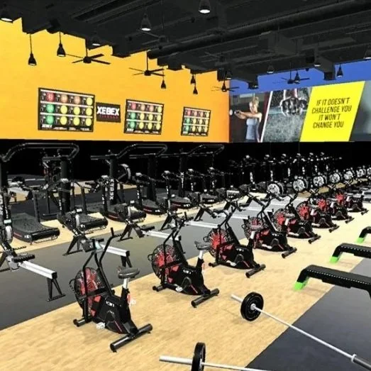 Indoor gym with cardio equipment, including stationary bikes, treadmills, and rowing machines, arranged in rows against a yellow and blue wall with motivational posters.