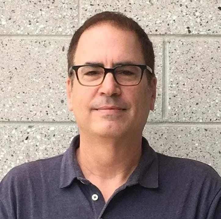 Man with short dark hair, wearing glasses and a dark collared shirt, standing in front of a gray textured wall.