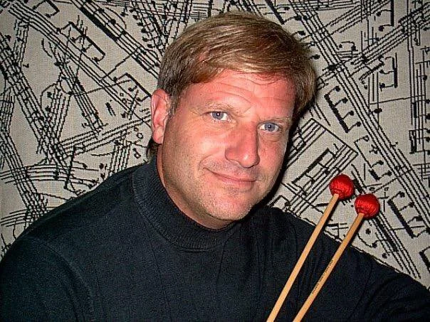 A man holding drumsticks with red tips, sitting in front of a background featuring a decorative musical score.