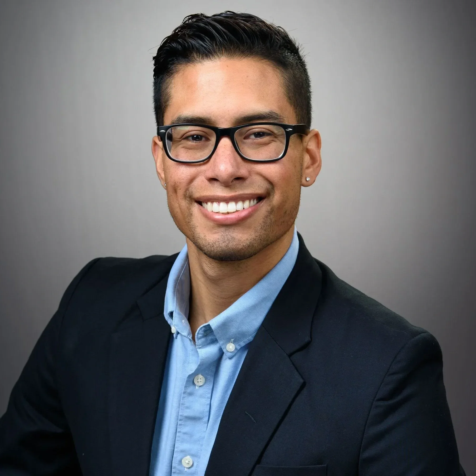 Professional headshot of a smiling man wearing glasses, a black blazer, and a light blue button-down shirt against a gray background.