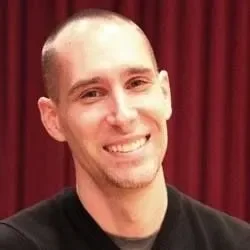 A young man with a shaved head smiling in front of a red curtain.