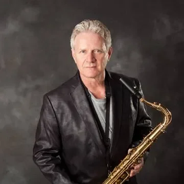 A man with gray hair wearing a black leather jacket holds a gold-colored saxophone against a dark gray background.