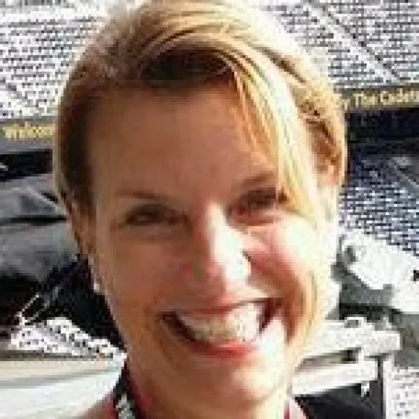 Close-up photo of a woman with short blonde hair smiling with teeth visible, background appears to be an outdoor setting with text on a screen or banner.