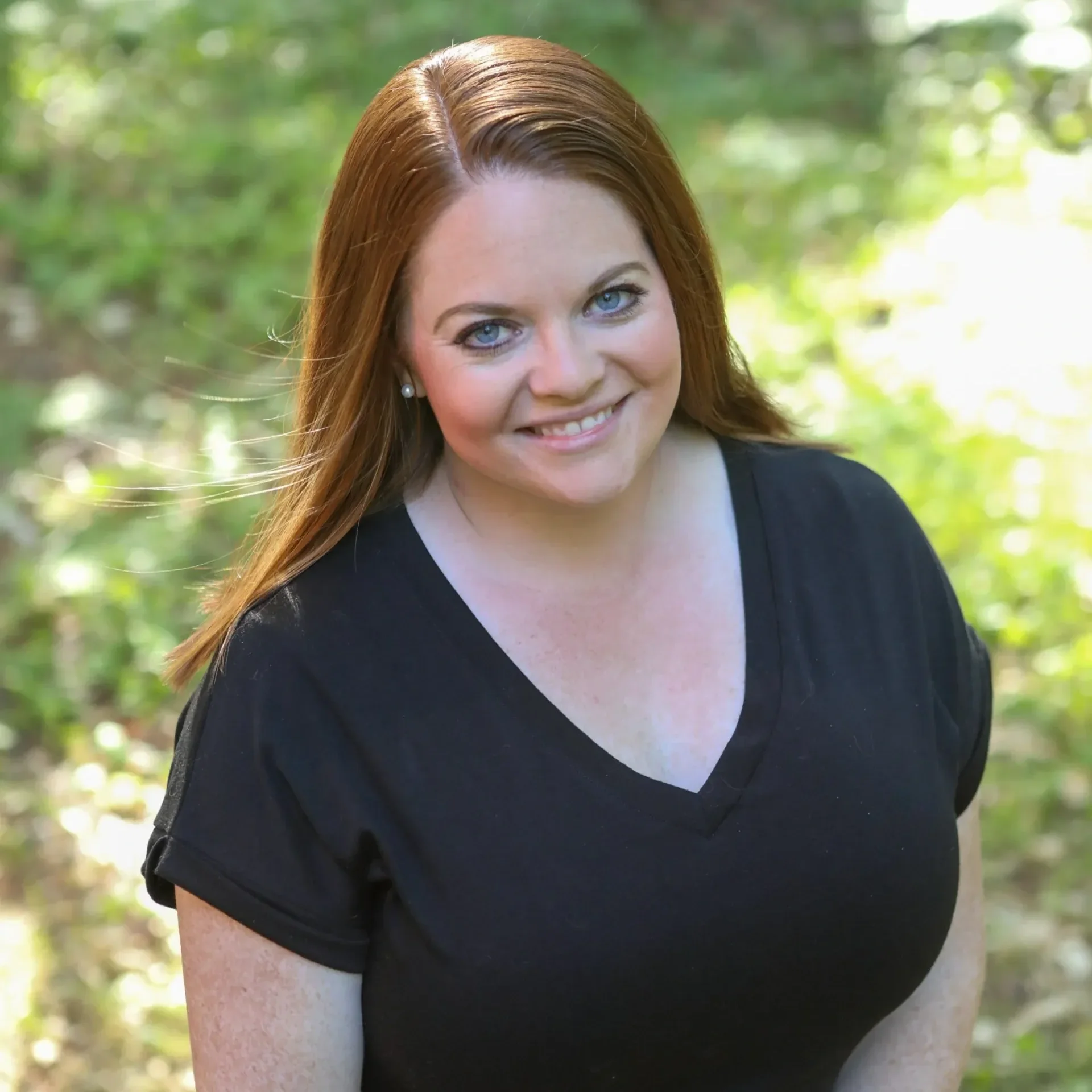 A woman with long red hair smiling outdoors with greenery in the background.