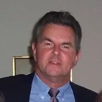 A man with dark hair and fair skin wearing a dark blazer, light blue shirt, and patterned tie, posing indoors.