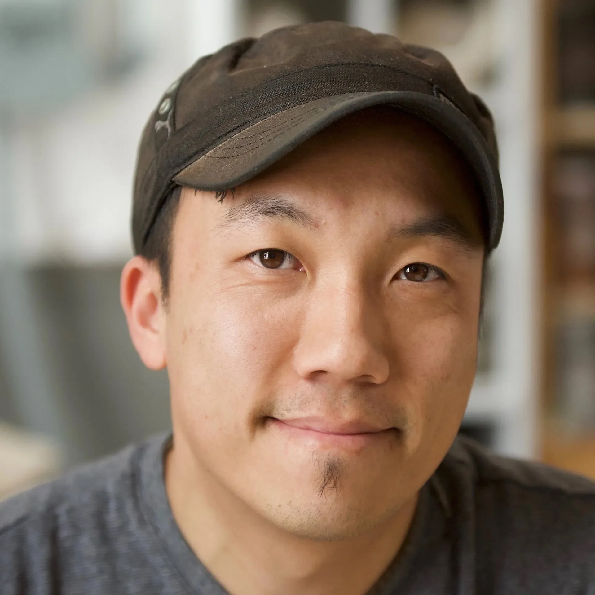Close-up of a smiling man wearing a brown cap, with a blurred indoor background.