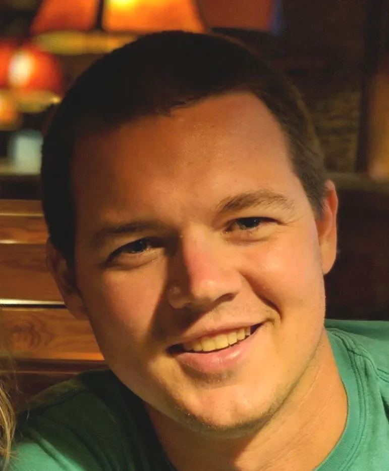 Close-up of a young man smiling with dark hair, wearing a green shirt, in a dimly lit environment with warm lighting.