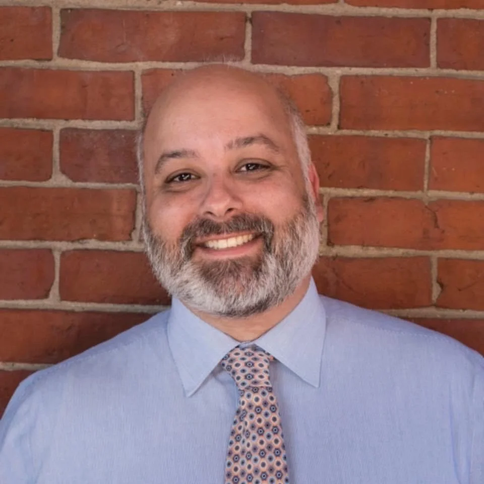A smiling man with a gray beard and short hair, wearing a light blue dress shirt and patterned tie, standing in front of a brick wall.