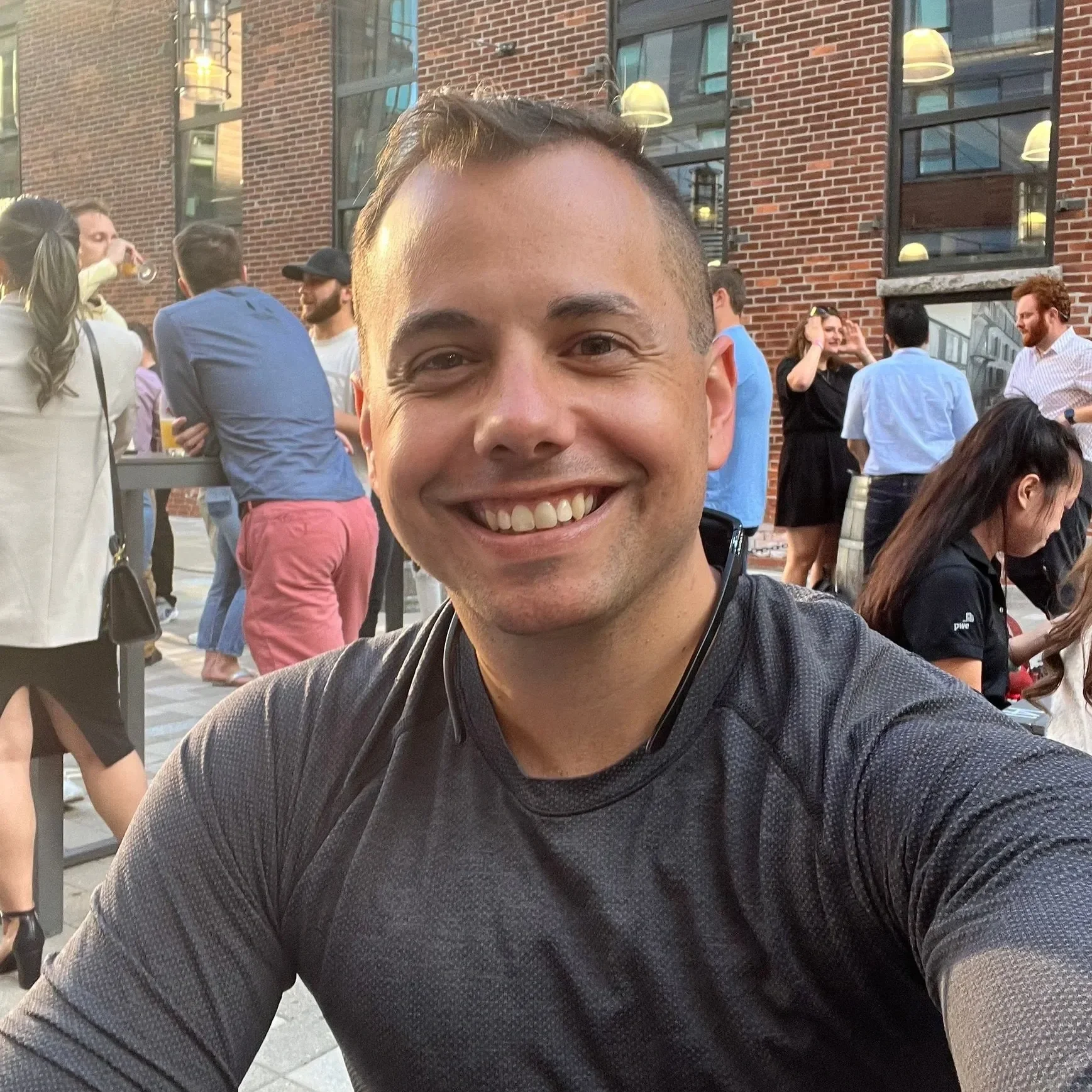 A smiling man taking a selfie at an outdoor social event with a brick building and group of people in the background.