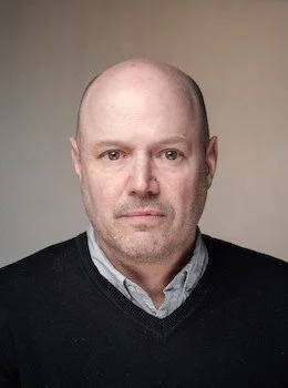A middle-aged man with a bald head and light complexion, wearing a black sweater over a collared shirt, looking directly at the camera against a plain background.