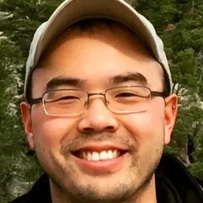 A smiling young man wearing glasses and a baseball cap outdoors with trees in the background.