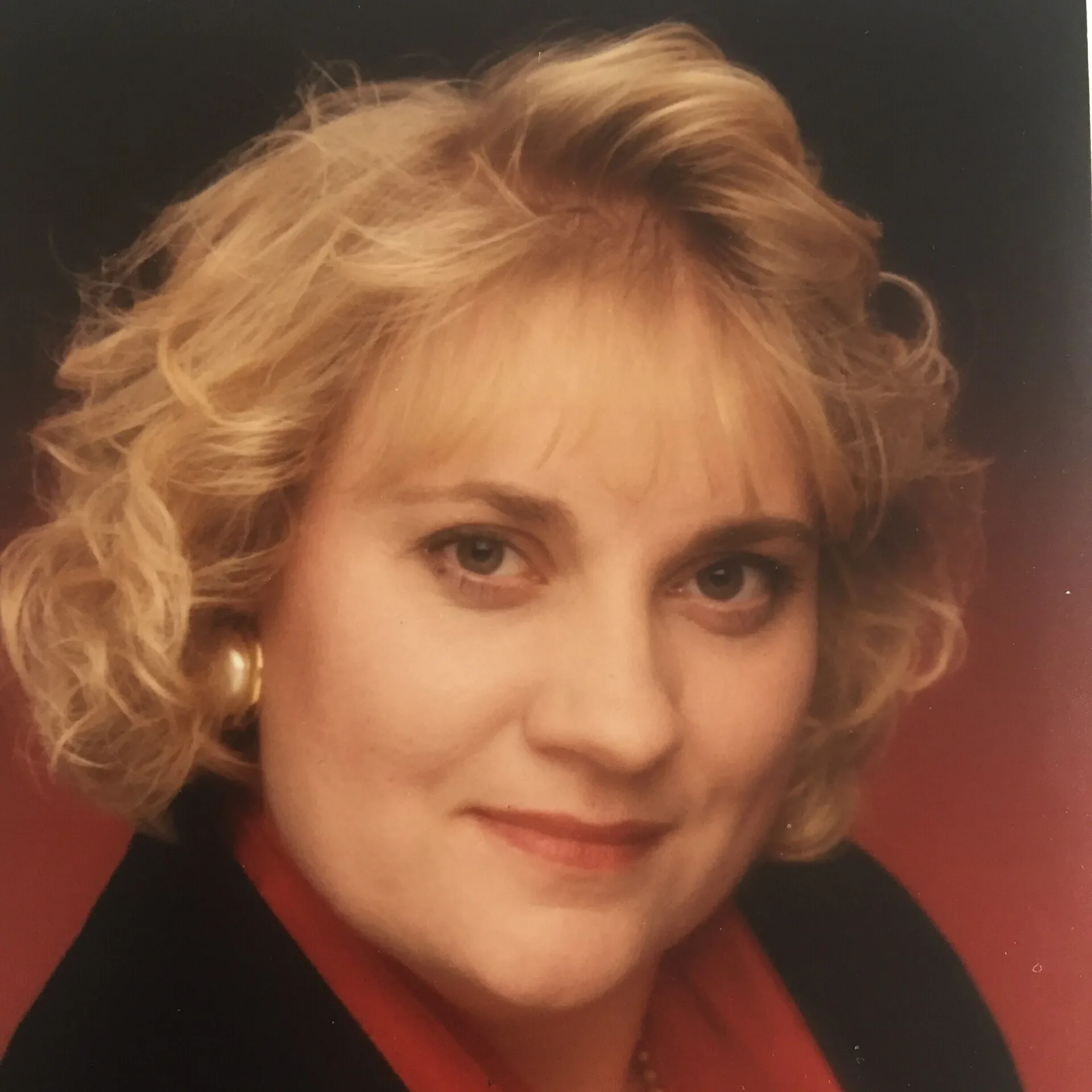 Close-up portrait of a woman with curly blonde hair, wearing a red top, gold earrings, and a black blazer, against a dark background.