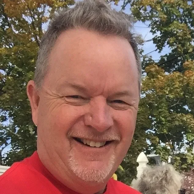 Smiling middle-aged man with short brown hair and a goatee, wearing a red shirt, outdoors with trees in the background.